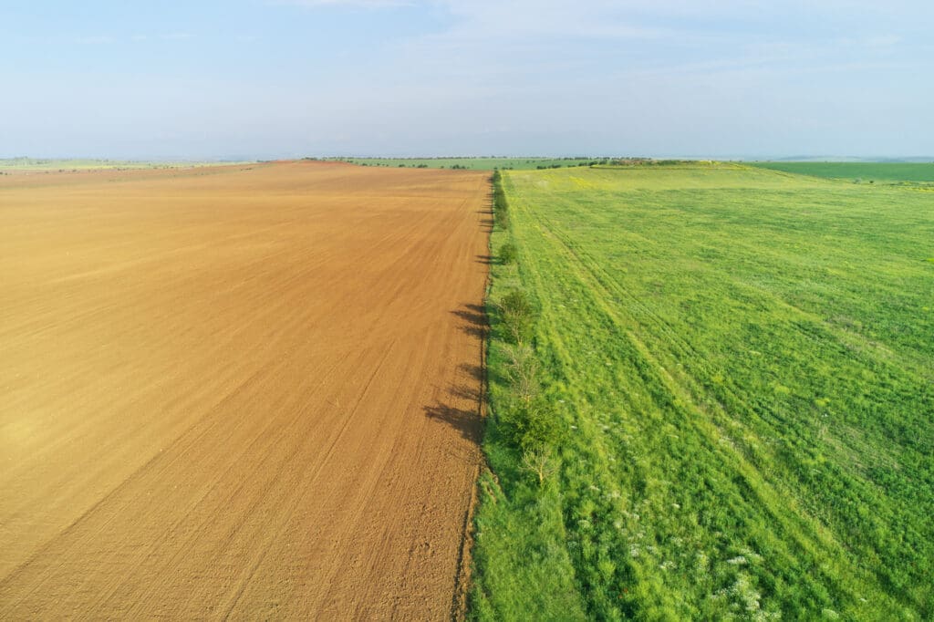 Aerial view of a landscape showcasing the benefits of regenerative agriculture, with a brown, plowed field on the left contrasting against a lush green field on the right, under a clear sky.
