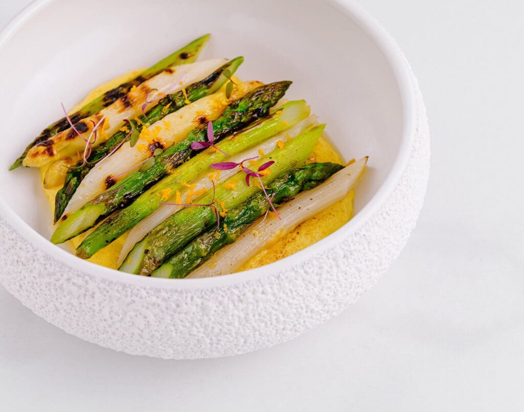 Grilled and roasted asparagus and white asparagus spears arranged neatly in a white textured bowl, garnished with microgreens, a hint of lemon, and a light sauce, on a white background.