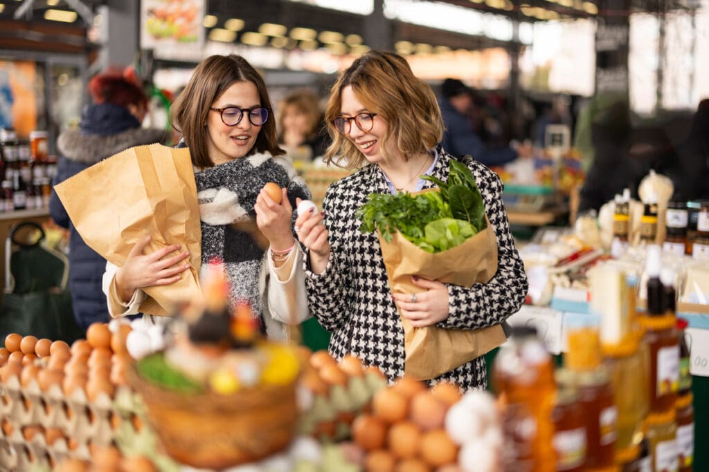 Two women shopping at a local farmers market with paper bags full of fresh organic produce, reducing their carbon footprint through sustainable food choices