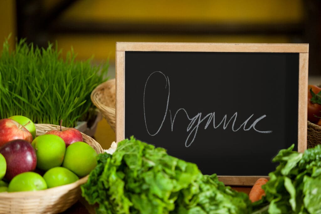 A blackboard with Organic written on it is surrounded by an assortment of fresh, natural produce, including green and red apples, leafy greens, and a pot of grass. The background softly blurs, emphasizing the wooden baskets and colorful fruits with handwritten labels.