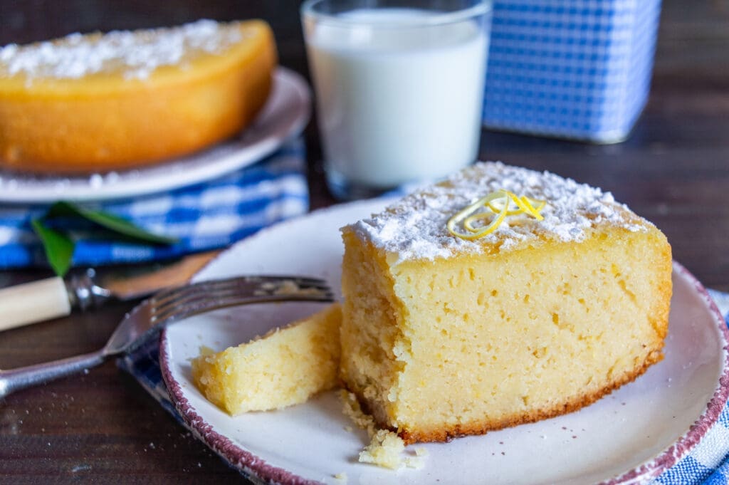 A slice of lemon cornmeal cake on a plate, dusted with powdered sugar and garnished with fresh lemon zest. A fork rests beside the cake. A glass of milk and another plate with cake sit in the background on a dark wooden table.