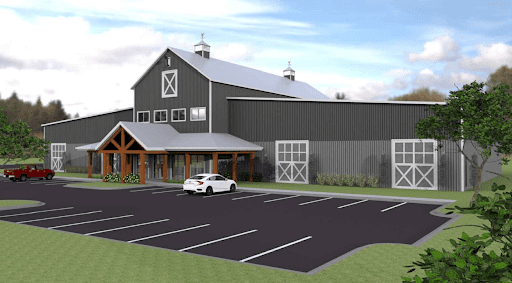 A modern barn-style building with gray metal siding, large barn doors, and wooden entryway accents, sits beside a parking lot with a few cars on a partly cloudy day. Green trees and grass surround the area.