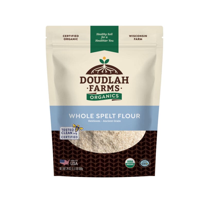 A pouch of Spelt Flour with a clear window displays the flour inside. The packaging, in blue and beige, showcases certifications such as USDA Organic and Non-GMO.
