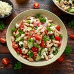 A bowl of cold salad with cranberry beans, cherry tomatoes, cucumbers, red onions, parsley, and crumbled feta cheese sits on a rustic wooden table, with extra feta and vegetables scattered around.