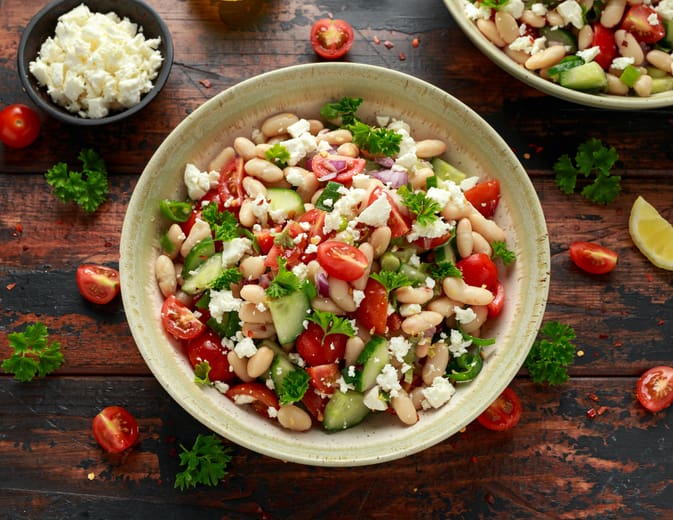 A bowl of cold salad with cranberry beans, cherry tomatoes, cucumbers, red onions, parsley, and crumbled feta cheese sits on a rustic wooden table, with extra feta and vegetables scattered around.