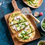 Flatbread topped with arugula, sliced radishes, zucchini ribbons, and dollops of cheese is cut into pieces on a wooden serving board. A pizza cutter, bowls of ingredients, and scattered greens surround the board.