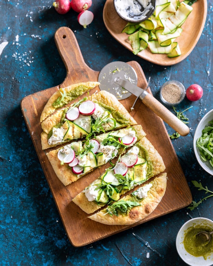 Flatbread topped with arugula, sliced radishes, zucchini ribbons, and dollops of cheese is cut into pieces on a wooden serving board. A pizza cutter, bowls of ingredients, and scattered greens surround the board.