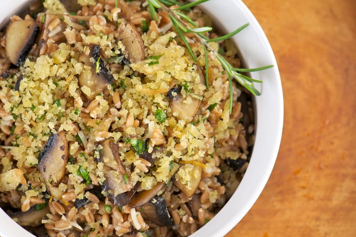 A close-up of a herb bowl of mushroom risotto featuring Emmer grain, topped with golden breadcrumbs and garnished with a sprig of rosemary, set on a wooden surface.
