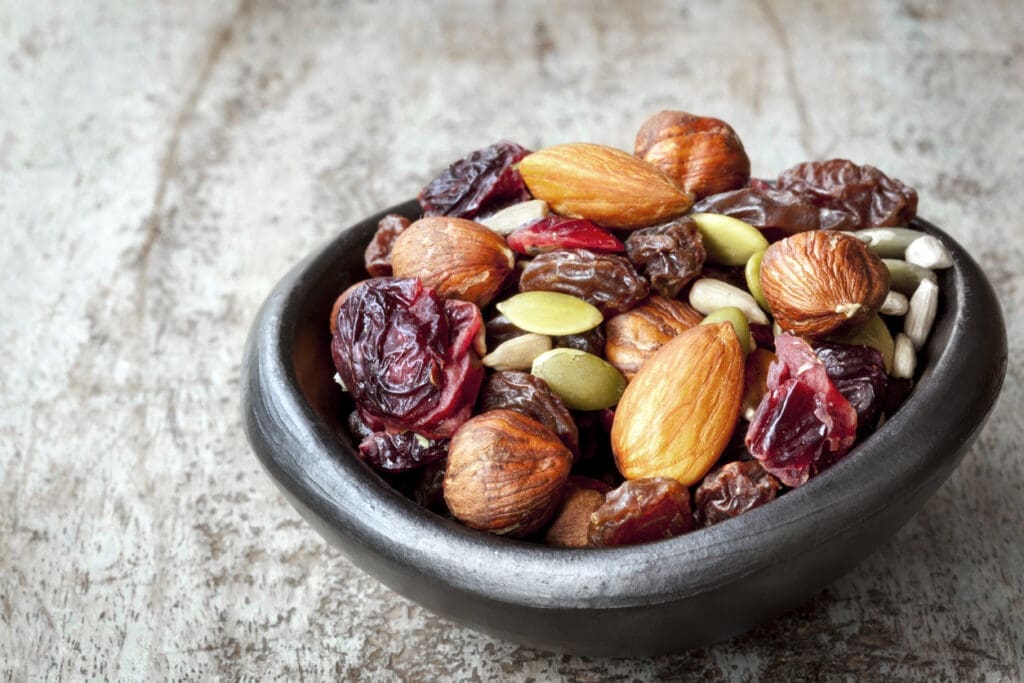 A small, dark bowl filled with mixed nuts, seeds, dried cranberries, and raisins sits on a rustic wooden surface.