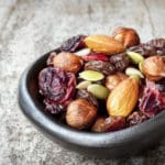 A small, dark bowl filled with mixed nuts, seeds, dried cranberries, and raisins sits on a rustic wooden surface.