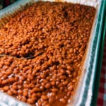 A large aluminum tray filled with baked beans in sauce sits on a red and white checkered tablecloth, with a metal serving spoon resting inside the tray.