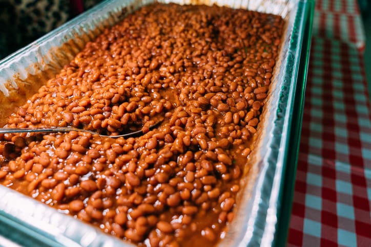 A large aluminum tray filled with baked beans in sauce sits on a red and white checkered tablecloth, with a metal serving spoon resting inside the tray.
