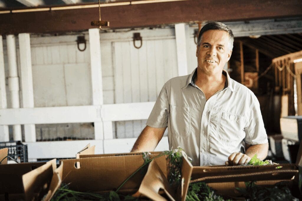 A man in a light shirt stands smiling behind boxes of freshly harvested Doudlah Farms Organics vegetables inside a rustic wooden barn. Sunlight filters in, creating a warm, inviting atmosphere.