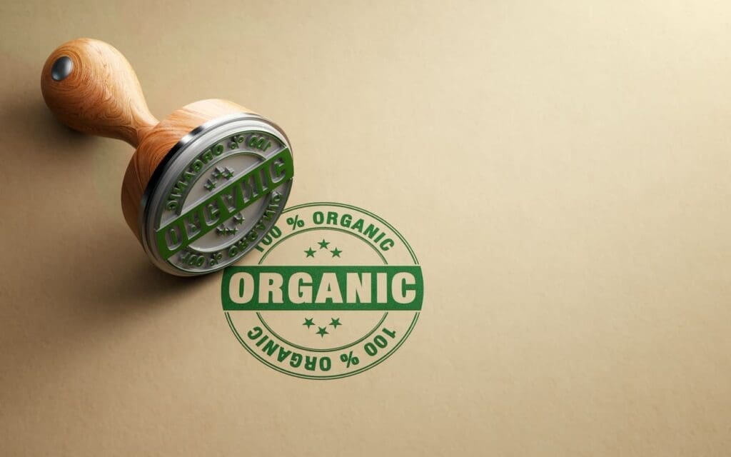 A wooden stamp with a metal base labeled ORGANIC rests on a brown surface, next to a green stamped mark that reads 100% ORGANIC in a circular design.