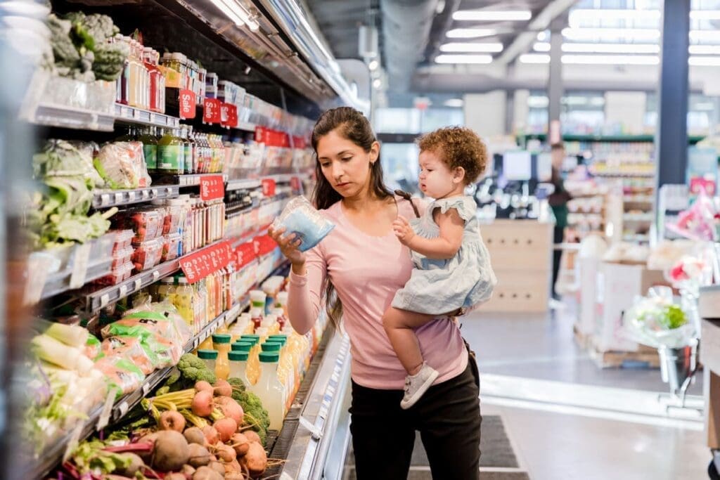 A woman holding a young child shops for groceries, examining a packaged item in the refrigerated produce section of a supermarket. Fresh vegetables and drinks are visible on the shelves.