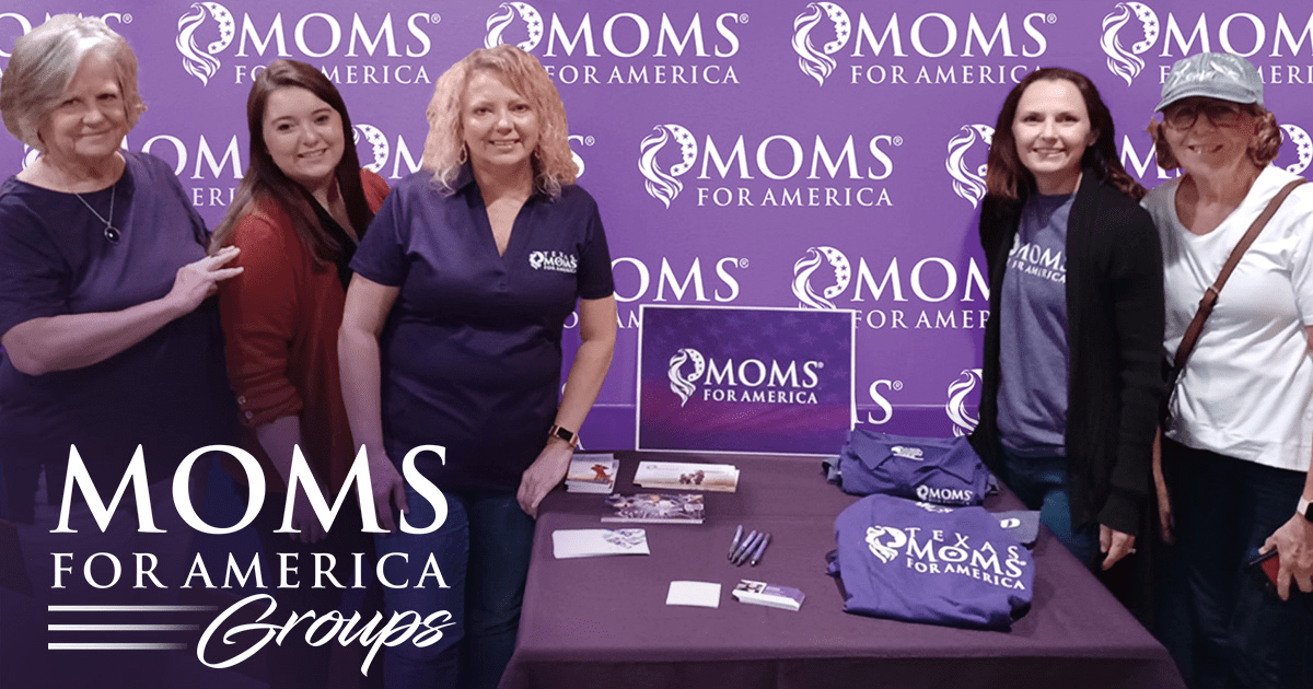 Five women stand and smile behind a table with Moms for America shirts, pens, and brochures—plus info on organic food from Doudlah Farms—in front of a purple Moms for America backdrop. “Moms for America Groups” appears in the lower left corner.