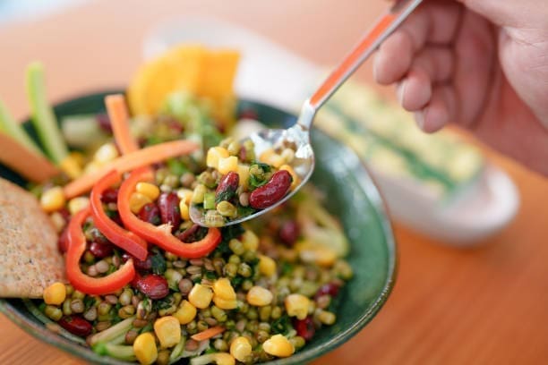 A hand holds a spoonful of colorful salad with kidney beans, corn, mung beans, and sliced red bell peppers above a bowl filled with the same ingredients. Crackers and vegetables are visible on the side.