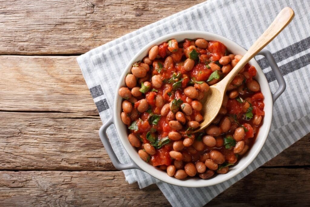 A white dish filled with stewed beans, tomatoes, and herbs sits on a gray-striped cloth atop a wooden surface; a wooden spoon rests inside the dish.