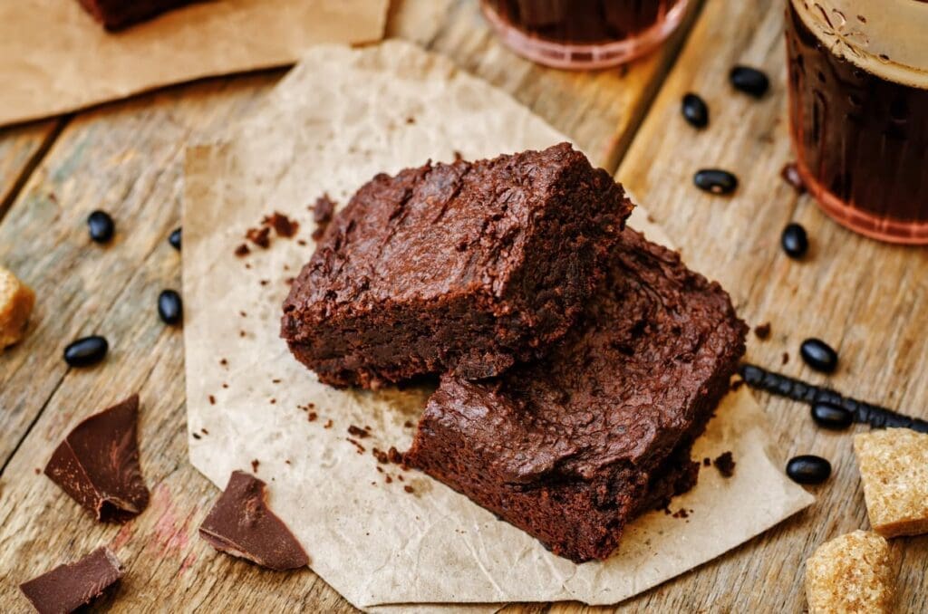 Two rich, fudgy chocolate brownies stacked on parchment paper, with coffee beans, sugar cubes, and broken chocolate pieces scattered on a rustic wooden table.