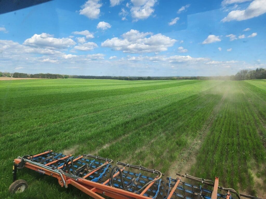A large green field under a blue sky with scattered clouds, with Doudlah Farms equipment in the foreground working the soil. Dust rises behind the machine as it moves through the crops, supporting organic food production.