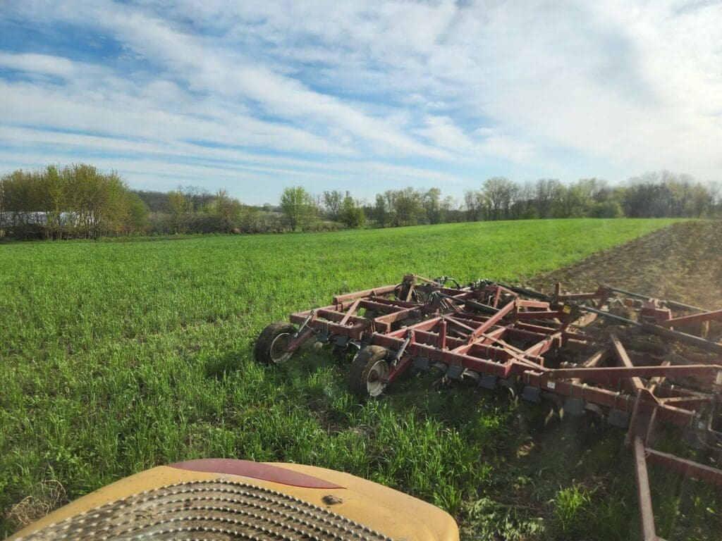 A large red agricultural harrow sits on a green field under a partly cloudy sky, with trees and distant hills in the background. The edge of farm equipment is visible in the foreground.