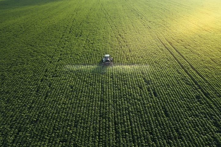 Aerial view of a tractor spraying crops in a large, lush green field under sunlight, with neat rows stretching into the distance.