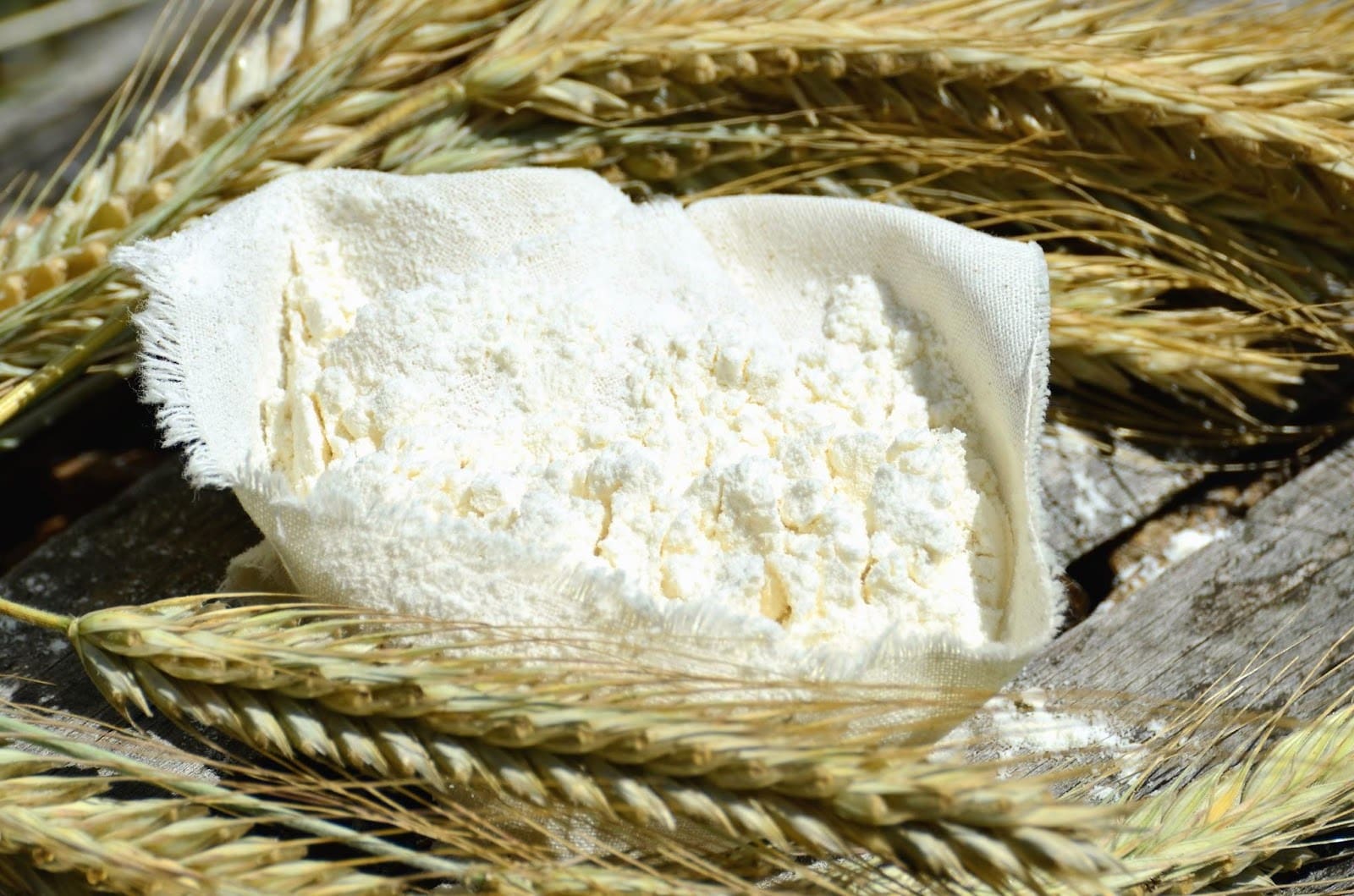 A small cloth pouch filled with stone ground flour is placed on a wooden surface, surrounded by golden wheat stalks.