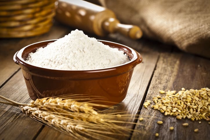 A brown ceramic bowl filled with organic flour sits on a wooden table next to wheat grains, wheat stalks, and a rolling pin. A burlap cloth and basket are in the background.