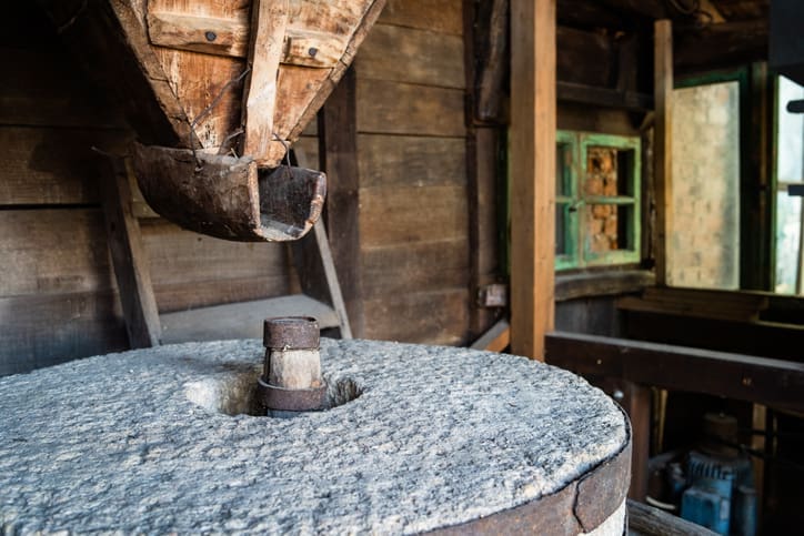 Close-up of an old stone mill inside a rustic wooden building, with a wooden chute above the grinding stone—showcasing the traditional process behind organic, stone ground flour. A small green window glows softly in the background.