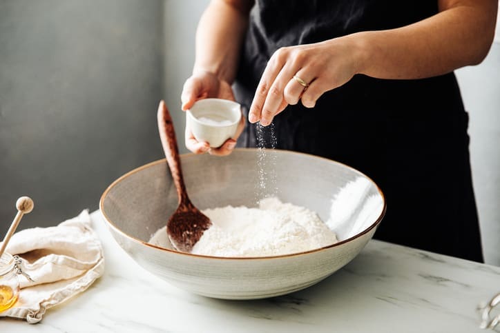 A person sprinkles salt into a large bowl of stone ground flour on a kitchen counter, preparing to bake. A wooden spoon rests in the bowl, and a beige cloth with utensils lies nearby.