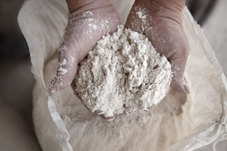 Close-up of two hands holding a heap of organic flour above an open sack, with flour dust visible on the hands and fabric.