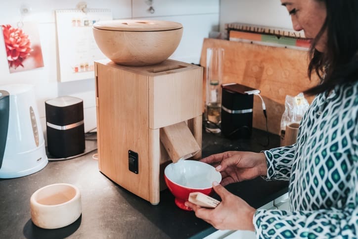 A woman stands in a kitchen, holding a bowl under a wooden grain mill as fresh stone ground flour dispenses into it. Various kitchen items and appliances are visible on the counter around her.