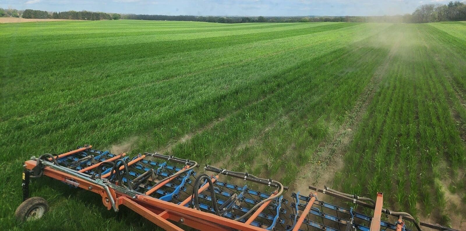 A large, orange agricultural harrow cultivates a green field under a blue sky, creating neat rows in the soil—an essential process in holistic agriculture that supports healthy, sustainable farmland.
