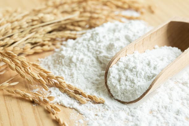 A wooden scoop filled with white rice flour rests on a wooden surface, surrounded by a pile of rice flour and stalks of unhulled rice.