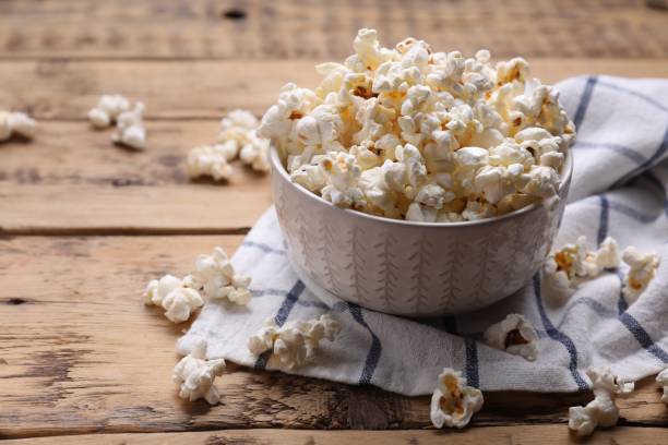 A white bowl filled with popcorn sits on a blue and white checkered cloth on a rustic wooden table, with some pieces of popcorn scattered around.