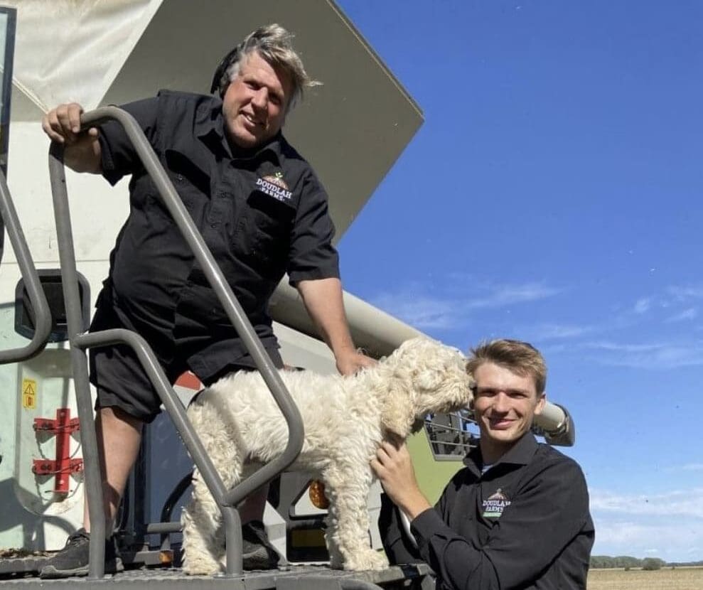 Two men in black shirts stand on metal steps beside Doudlah Farms Organics equipment, smiling at the camera. One stands and pets a fluffy white dog, while the other kneels to join in. The sky is clear and blue, highlighting their passion for organics.