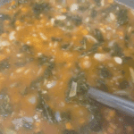 A close-up of a pot filled with vegetable soup featuring leafy greens, white beans, diced carrots, onions, and a tomato-based broth. A metal ladle is partially visible, stirring the ingredients.