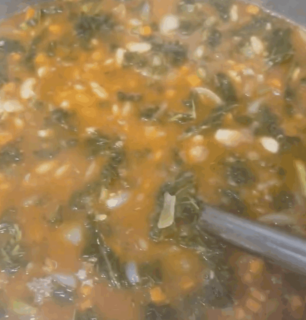 A close-up of a pot filled with vegetable soup featuring leafy greens, white beans, diced carrots, onions, and a tomato-based broth. A metal ladle is partially visible, stirring the ingredients.