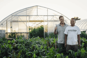 Two men stand smiling in front of a greenhouse, surrounded by lush green plants. One holds a garden tool, while the other wears a Farms2Food T-shirt. The scene suggests teamwork in a thriving vegetable garden.