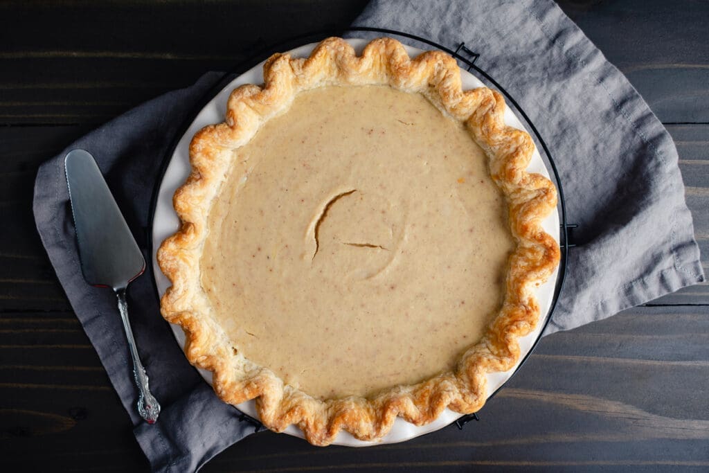 A whole pie with a golden, crimped crust and a smooth, light brown filling sits on a gray cloth atop a dark wooden table, with a pie server placed to the side.