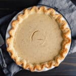 A whole pie with a golden, crimped crust and a smooth, light brown filling sits on a gray cloth atop a dark wooden table, with a pie server placed to the side.