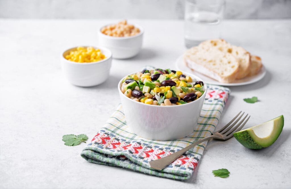 A white bowl filled with salad made of corn, black beans, and diced vegetables sits on a patterned napkin. Nearby are bowls of corn and grains, sliced bread, a glass of water, and a wedge of avocado.