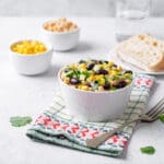 A white bowl filled with salad made of corn, black beans, and diced vegetables sits on a patterned napkin. Nearby are bowls of corn and grains, sliced bread, a glass of water, and a wedge of avocado.