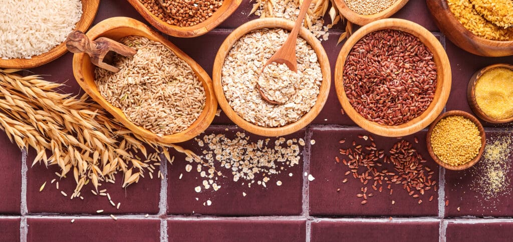 Various grains and cereals, including ancient grains and modern wheat, are displayed in wooden bowls on a red tiled surface, accompanied by a wooden scoop and a bundle of oat stalks. Notice the difference in textures and colors among them.