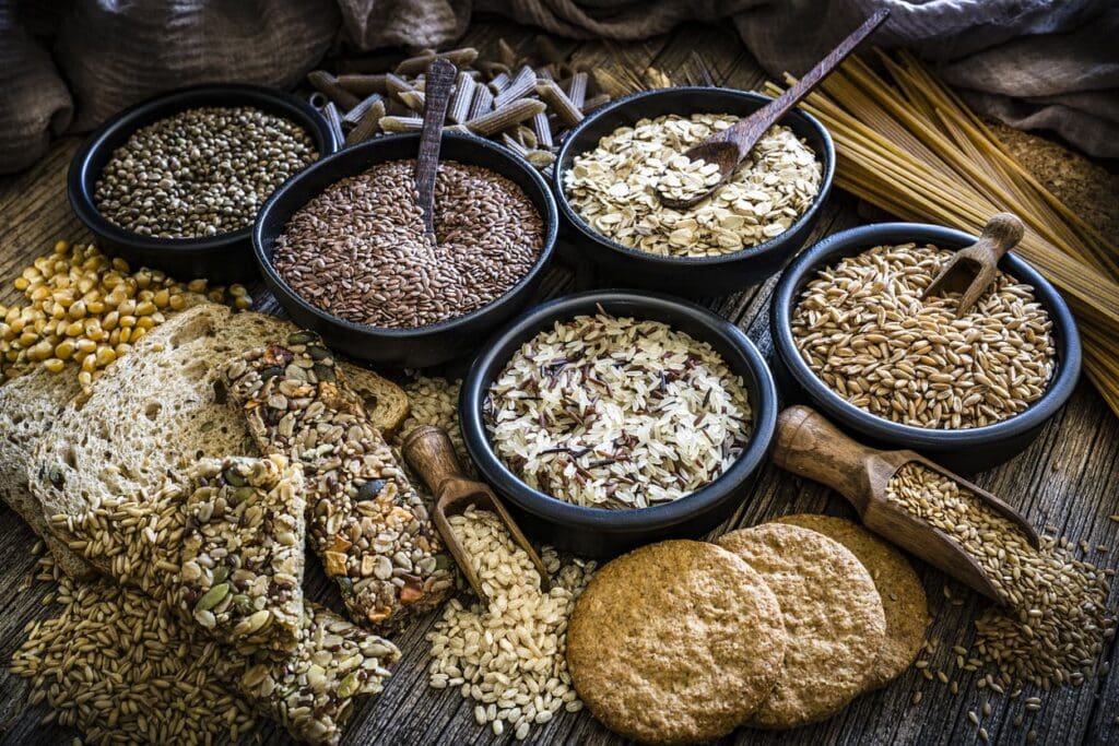 An assortment of ancient grains, seeds, rice, and oats in bowls, with slices of multigrain bread, crispbread, pasta, cookies, and loose grains displayed on a rustic wooden table highlights the difference from modern wheat varieties.
