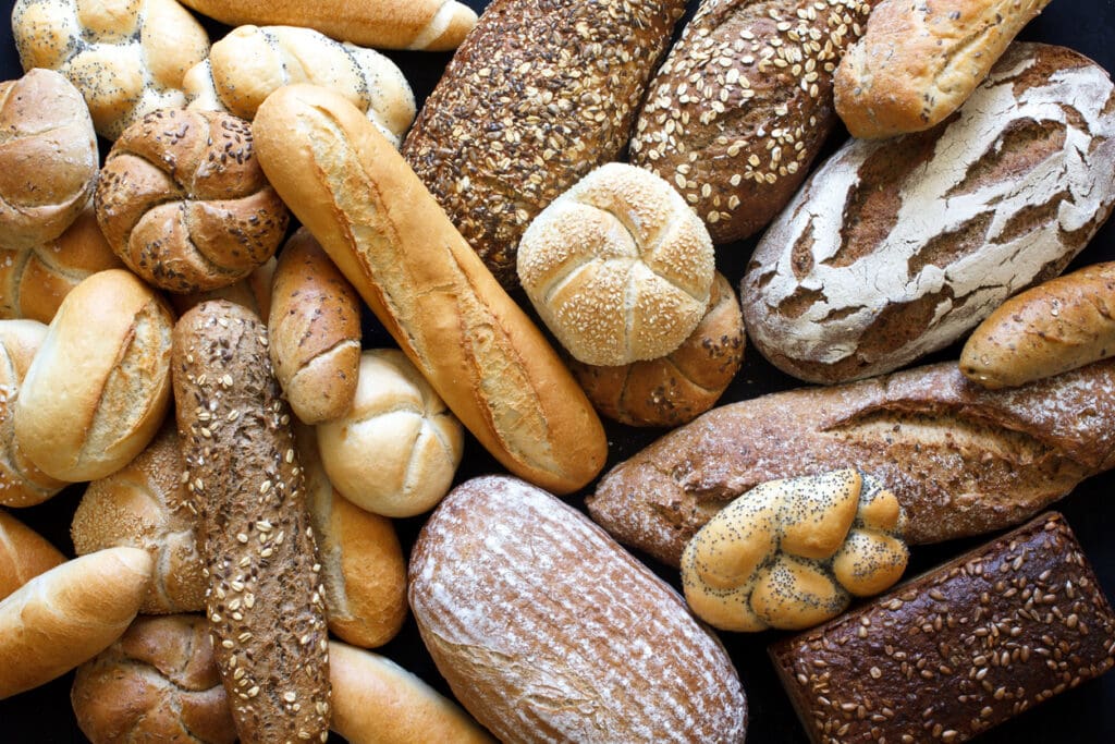 Assorted breads including baguettes, rolls, seeded loaves with ancient grains, round breads, and rustic artisan bread are arranged closely together, showcasing a difference in crusts, shapes, and grains.