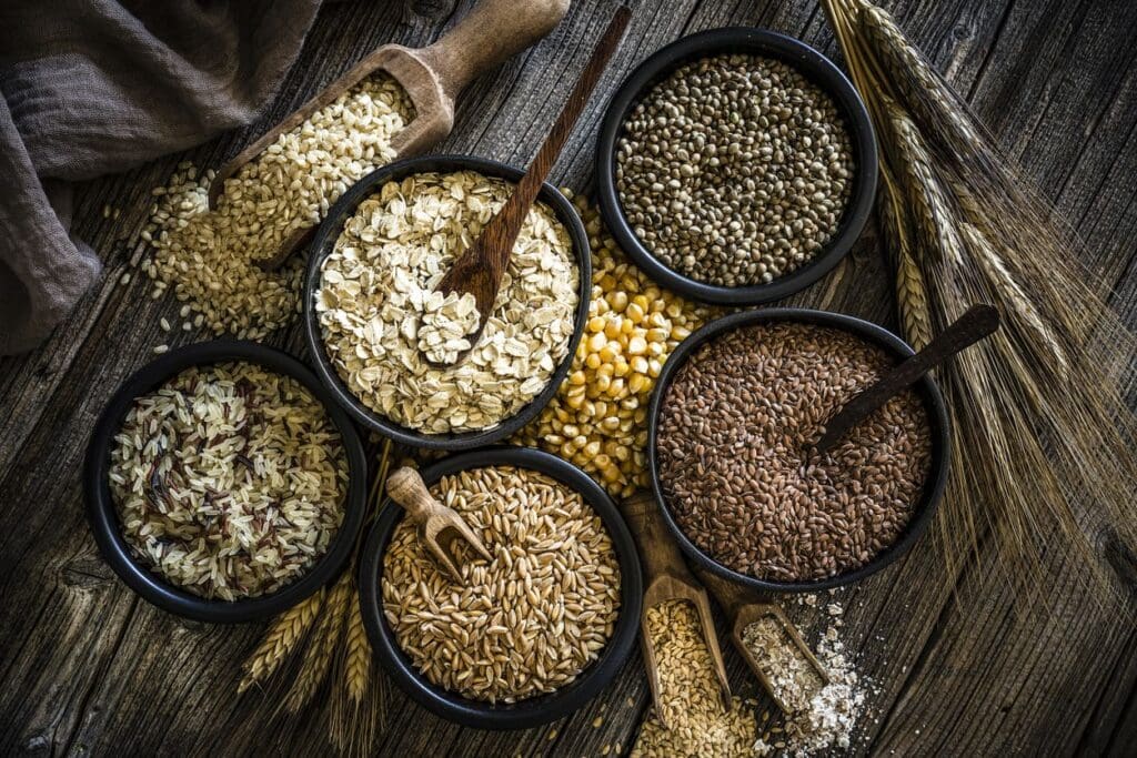 Assorted whole grains, including ancient grains like barley and millet, plus oats, brown rice, corn, and flaxseeds, displayed in black bowls on a rustic wooden table with scoops and stalks of modern wheat nearby.