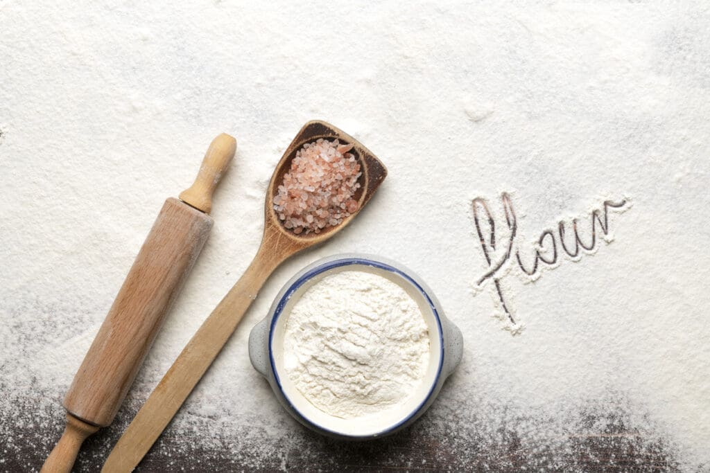 A rolling pin, a wooden spoon with pink salt, and a bowl of flour—made from ancient grains—sit on a floured surface. The word flour is written in the flour, highlighting the difference from modern wheat.