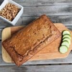 A loaf of zucchini bread rests on a wooden cutting board beside four cucumber slices. A small white bowl of chopped walnuts sits nearby on a rustic wooden table.