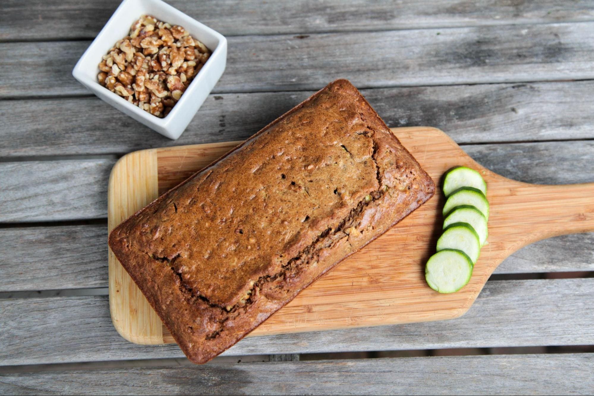 A loaf of zucchini bread rests on a wooden cutting board beside four cucumber slices. A small white bowl of chopped walnuts sits nearby on a rustic wooden table.